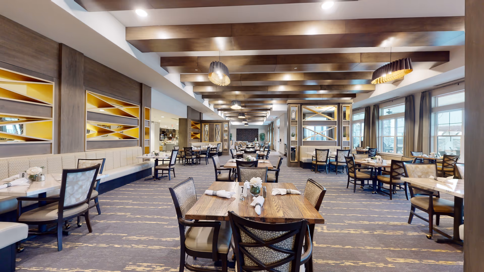 Spacious and well-lit dining room in a senior living facility with multiple wooden tables and chairs arranged neatly. The room features large windows with curtains, decorative ceiling beams, modern pendant lights, and built-in wall shelves with yellow accents. Tables are set with white napkins and small flower arrangements.