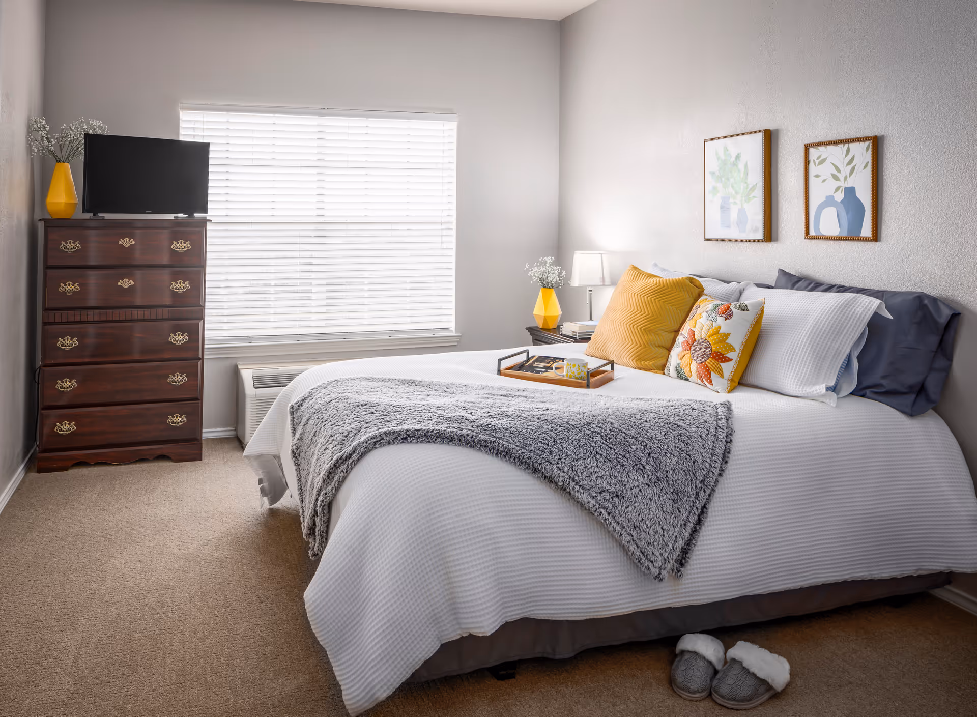 Bright, neatly staged bedroom with a made bed topped with decorative pillows and a throw, a wooden chest of drawers with a TV by a window, and slippers on the floor.