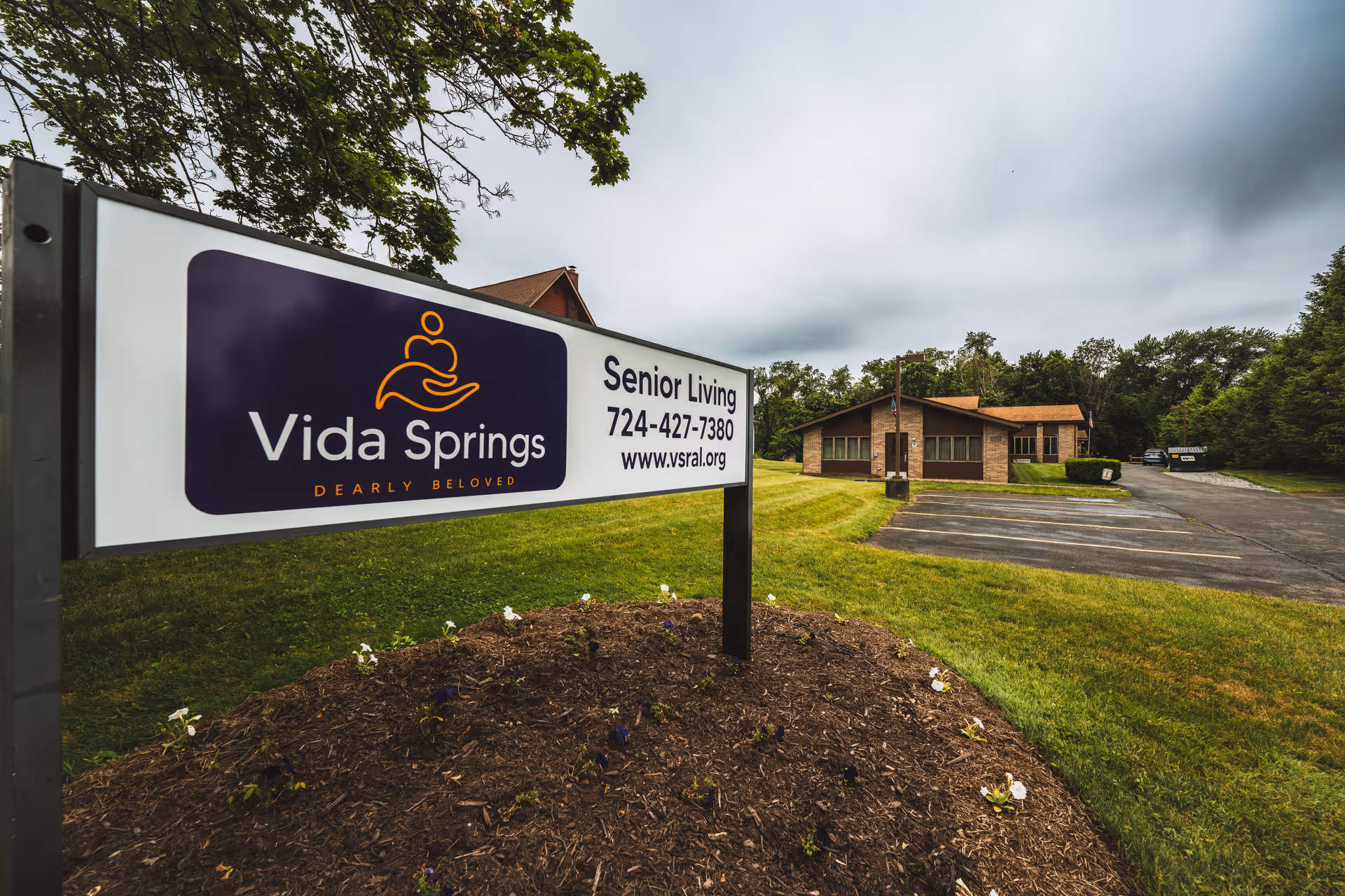 A Vida Springs Senior Living sign stands in front of a single-story brick building, parking lot, and grassy grounds under a cloudy sky.