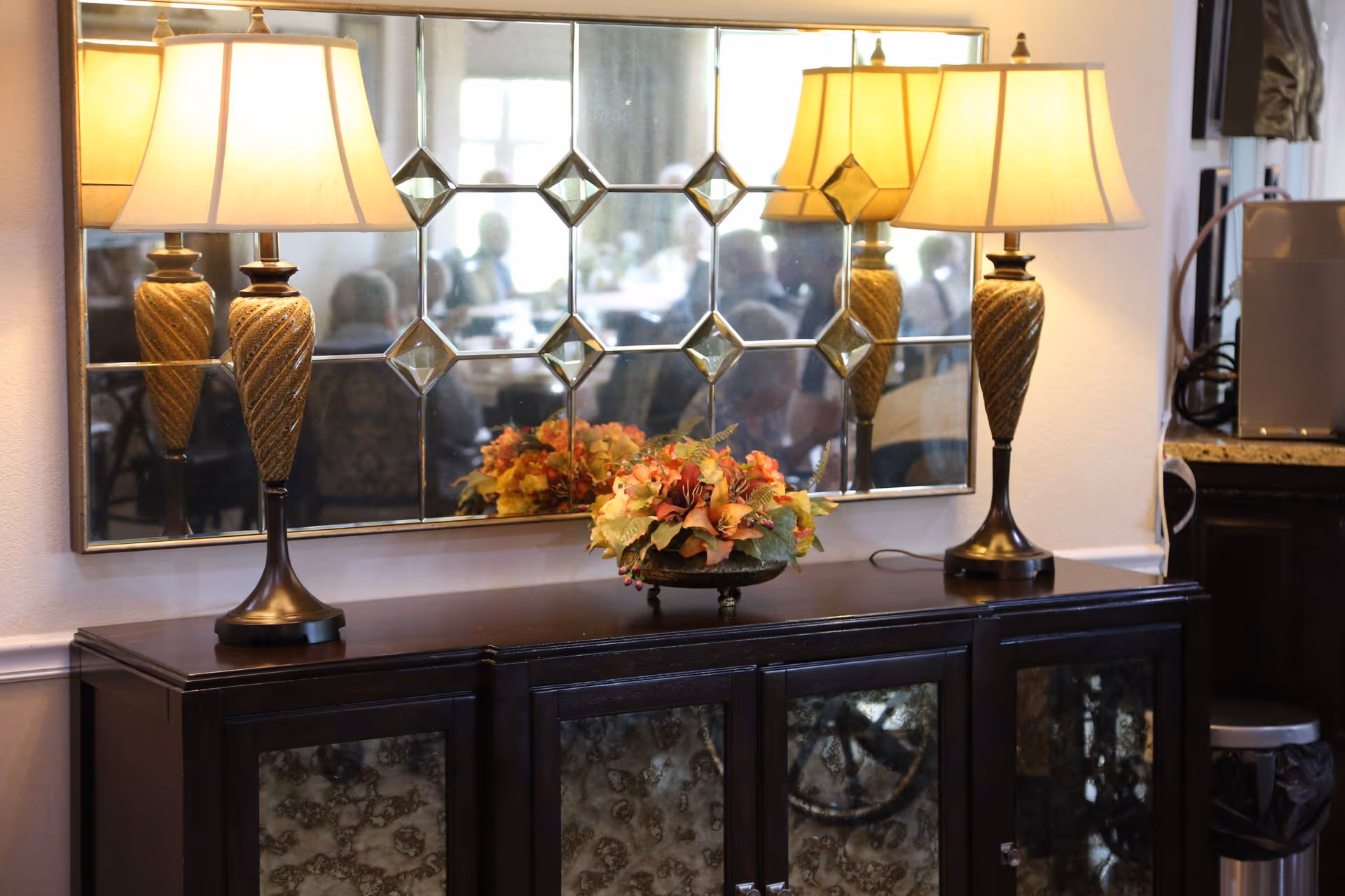 A dark wooden cabinet with glass-paneled doors topped with a decorative floral arrangement and two ornate table lamps with beige lampshades. Behind the cabinet is a large decorative mirror with a geometric pattern reflecting a blurred view of people sitting at tables.