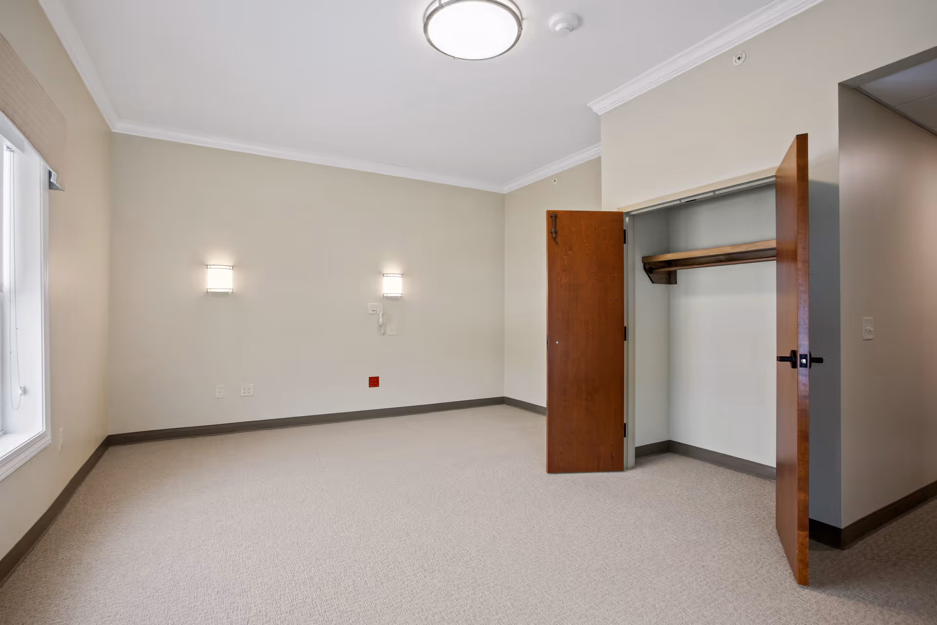 Empty senior living bedroom with beige walls, carpeted floor, two wall-mounted lights, a window with a shade, and an open closet with wooden doors and a hanging rod.