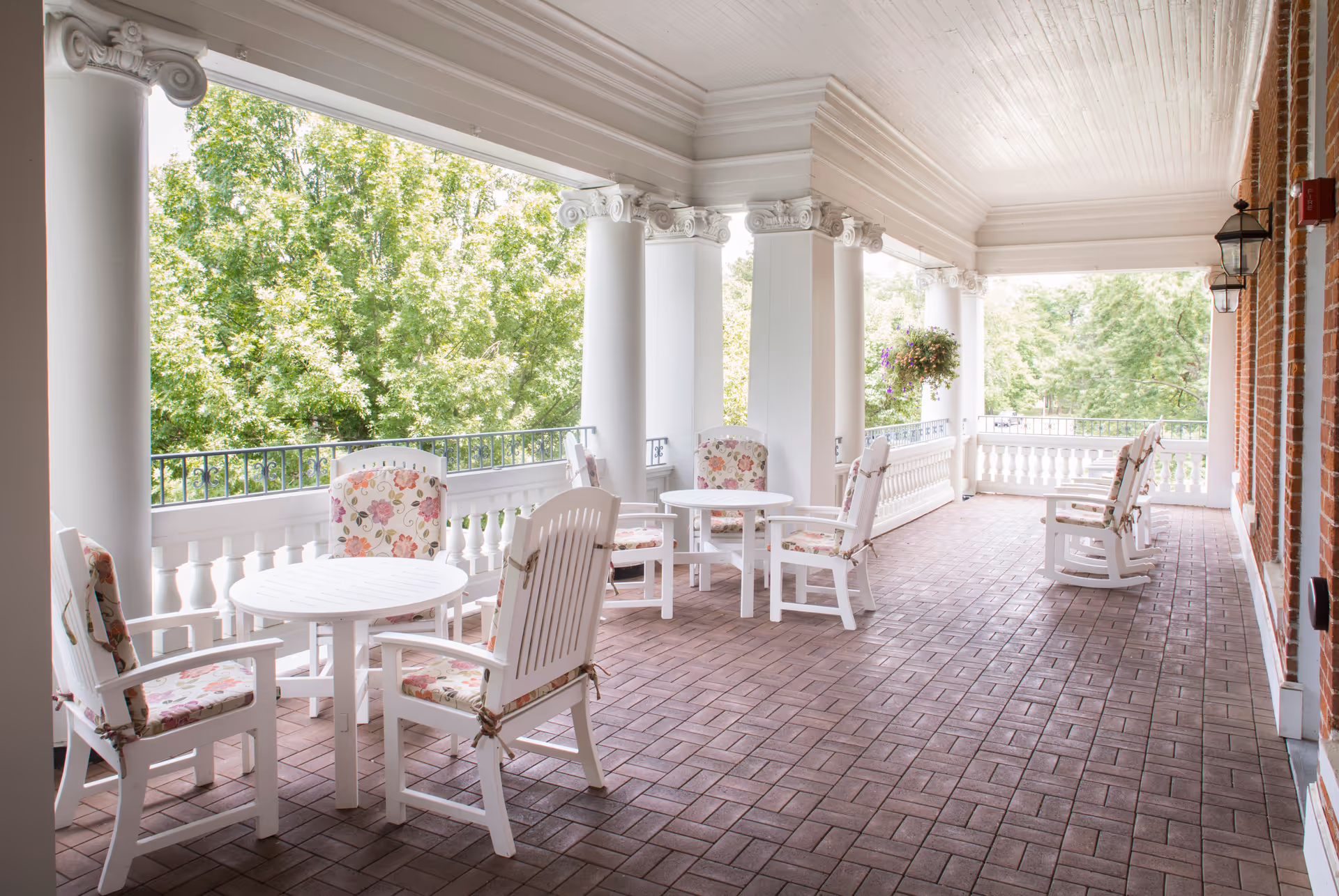 A spacious covered outdoor porch area with white columns and a brick floor. The porch is furnished with white wooden chairs and round tables, some chairs have floral cushions. Green trees are visible beyond the railing, and hanging flower baskets add to the decor.