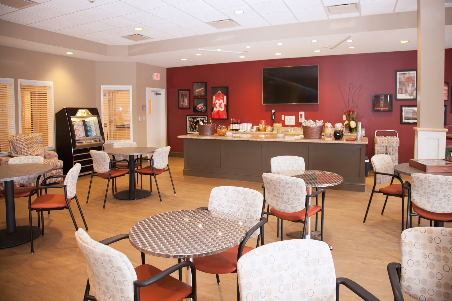 A spacious common area with several round tables and chairs arranged for seating. The room features a counter with various refreshments and snacks, a large flat-screen TV mounted on a red accent wall, framed sports memorabilia, and a jukebox in the corner. The flooring is wood, and the lighting is bright with recessed ceiling lights.