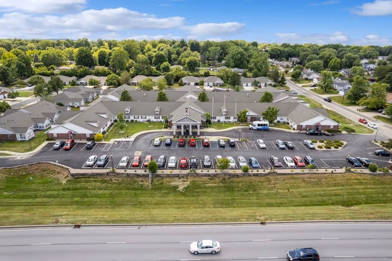 Aerial view of Five Star Residences of Banta Pointe, showing a large single-story building with a parking lot filled with cars in front. The building is surrounded by trees and residential houses, with a road running in front of the property.