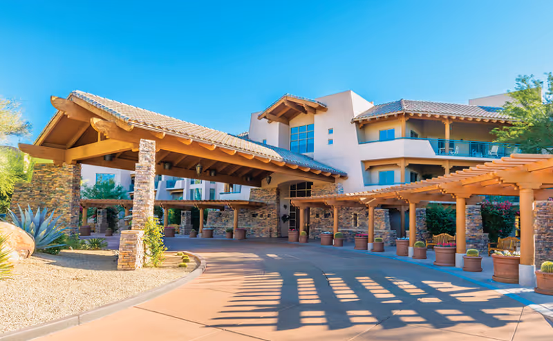 Exterior view of Vi at Grayhawk senior living facility featuring a covered entrance with stone pillars and wooden beams, a curved driveway, potted plants, and a multi-story building with balconies under a clear blue sky.