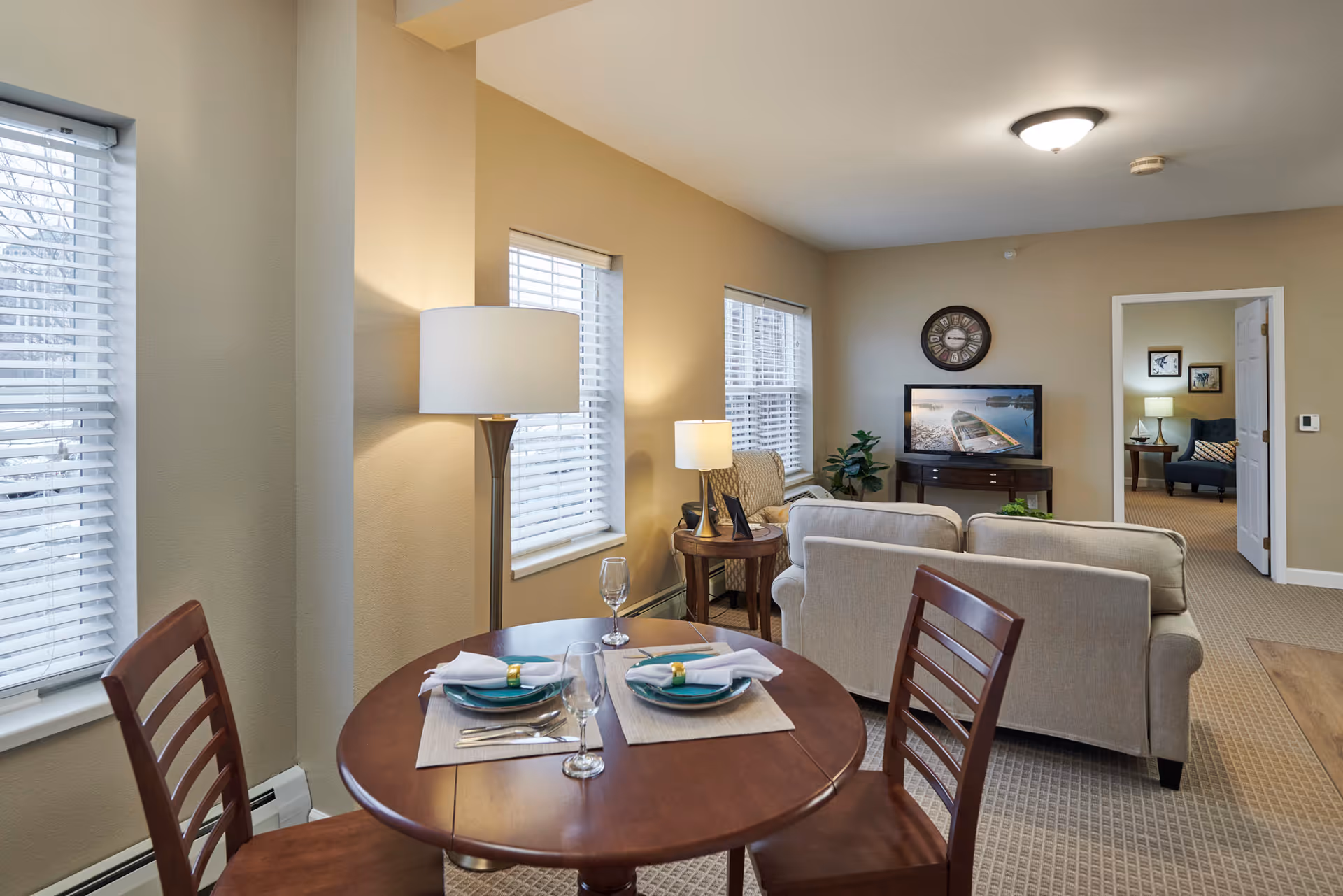 A cozy senior living area featuring a small round dining table set for two with plates, napkins, and glasses. Behind the table is a living room with a beige sofa, an armchair, a TV on a stand displaying an image of a boat on water, a wall clock above the TV, and a side table with a lamp. The room has beige walls, carpeted floors, and multiple windows with white blinds letting in natural light. An open doorway leads to another room with additional seating and framed artwork on the walls.