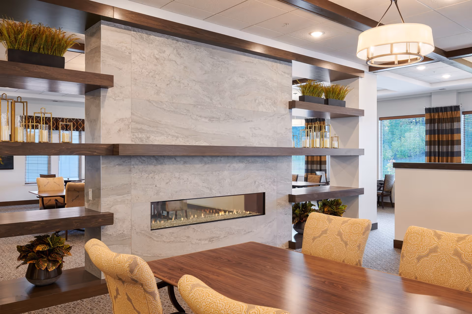 A cozy interior dining area with a wooden table and upholstered chairs featuring a yellow patterned fabric. A modern rectangular gas fireplace is built into a marble wall with dark wooden shelves on either side, decorated with plants and candle holders. Large windows with striped curtains allow natural light to fill the space.
