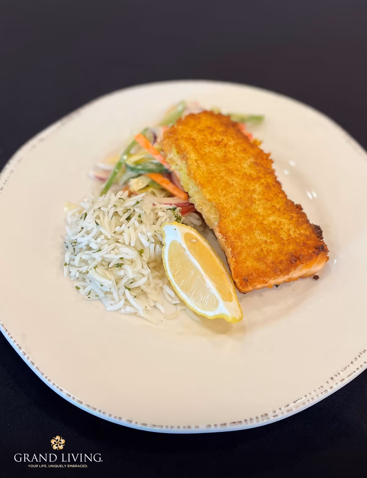A plate with a serving of breaded and fried fish fillet, a portion of white rice garnished with herbs, a lemon wedge, and a side of mixed vegetables including carrots and green beans.