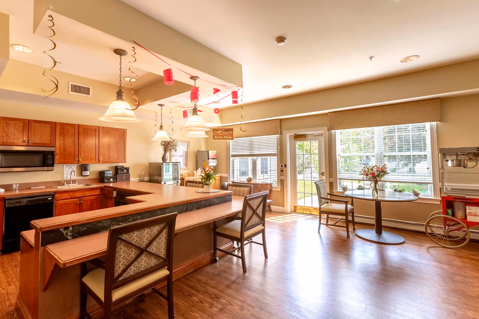 A bright and spacious kitchen and dining area with wooden cabinets, a microwave, and a dishwasher. There is a large counter with chairs for seating, pendant lights hanging from the ceiling, and festive red and gold hanging decorations. A small round table with two chairs is placed near large windows letting in natural light. A sign reading 'Hearth Bake Shop' is visible above a small counter area with a beverage dispenser and a popcorn machine on a red cart.