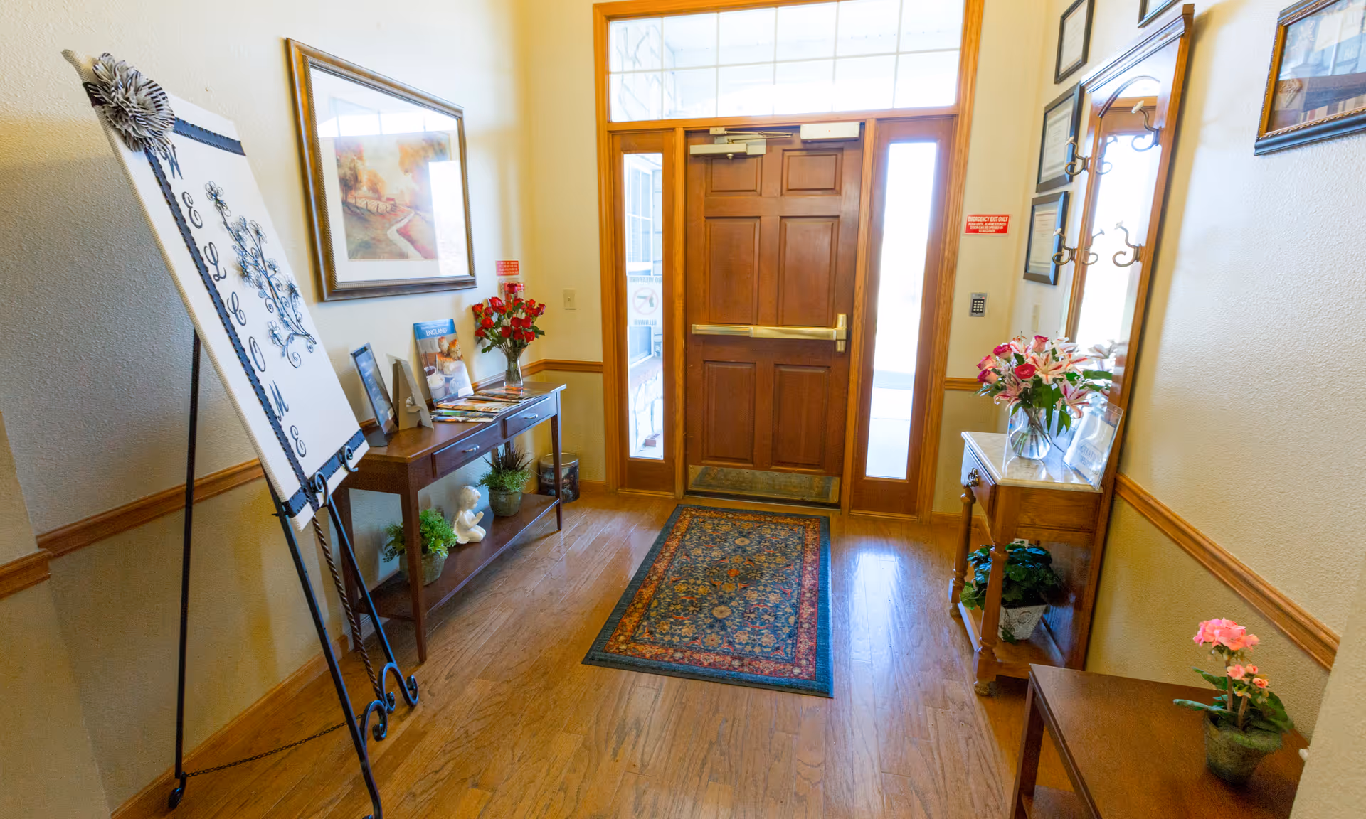 Well-lit entryway with a wooden front door, patterned rug, console tables with flowers and decor, and a welcome sign on an easel.