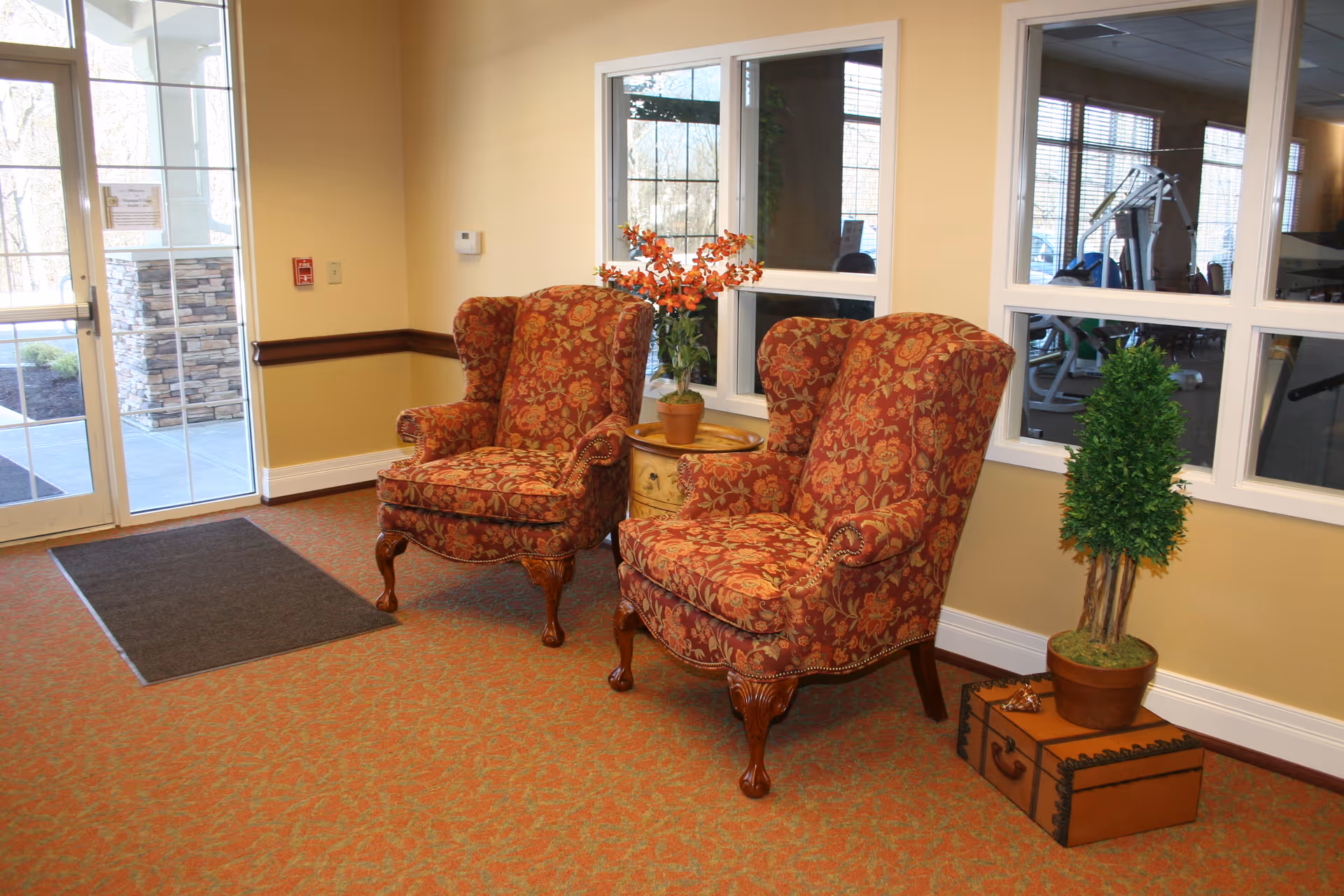 Two patterned upholstered armchairs with wooden legs are placed side by side in a well-lit room with beige walls and a patterned carpet. Between the chairs is a small round table with a potted plant featuring orange flowers. To the right of the chairs is a potted green plant on top of a decorative trunk. Large windows behind the chairs reveal a gym area with exercise equipment. A glass door to the left leads outside.