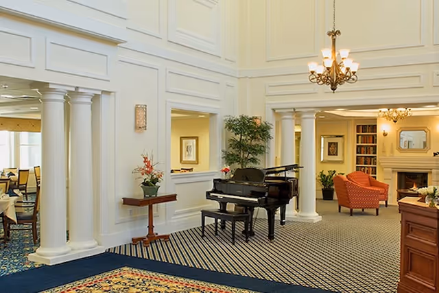 A spacious and elegant interior common area featuring a black grand piano with a bench, white columns, a chandelier, a patterned carpet, a small table with a flower arrangement, and a seating area with an orange armchair near a fireplace and bookshelves.