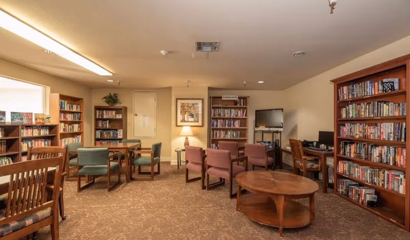 A cozy senior living common room with multiple wooden bookshelves filled with books, several wooden tables and chairs arranged for group seating, a floor lamp, a framed picture on the wall, and a TV on a stand in the corner.