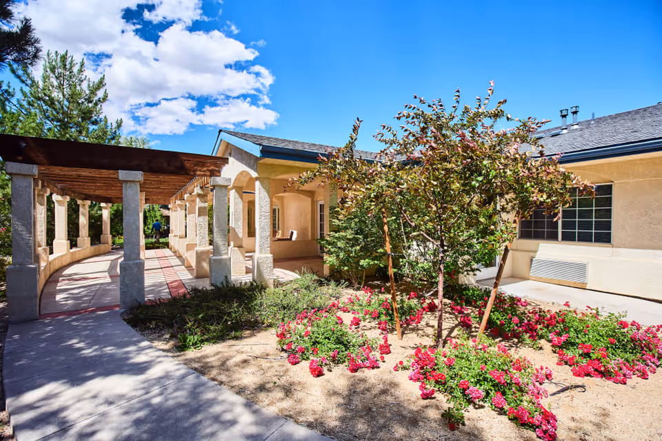 Sunlit courtyard with a pergola-covered walkway, flowering beds, and the beige exterior of a memory care building.