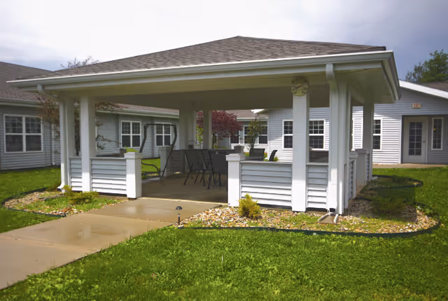 Covered outdoor pavilion with chairs and a table in a grassy courtyard surrounded by single-story white-siding buildings.