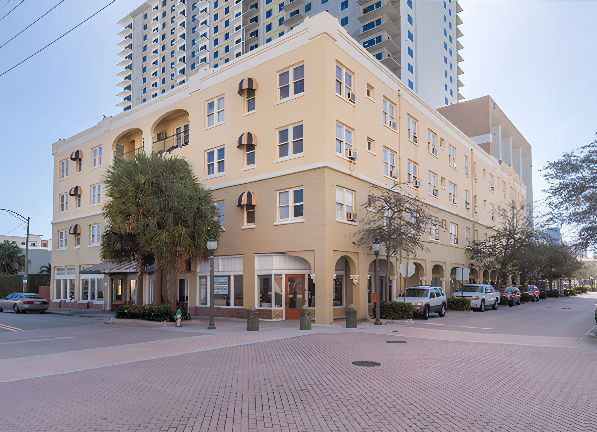 Exterior view of a multi-story beige building with several windows and small awnings above some windows. There are palm trees and other trees along the sidewalks, with cars parked on the street beside the building. The building is located at a street intersection with a brick-paved crosswalk.