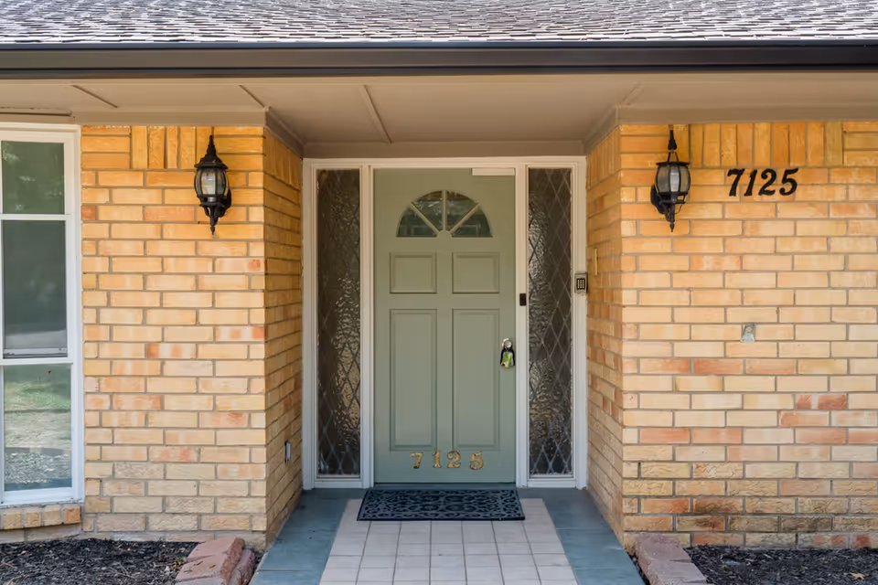 Brick exterior of a building with a green front door, sidelights, and lantern-style lights on either side of the entrance.