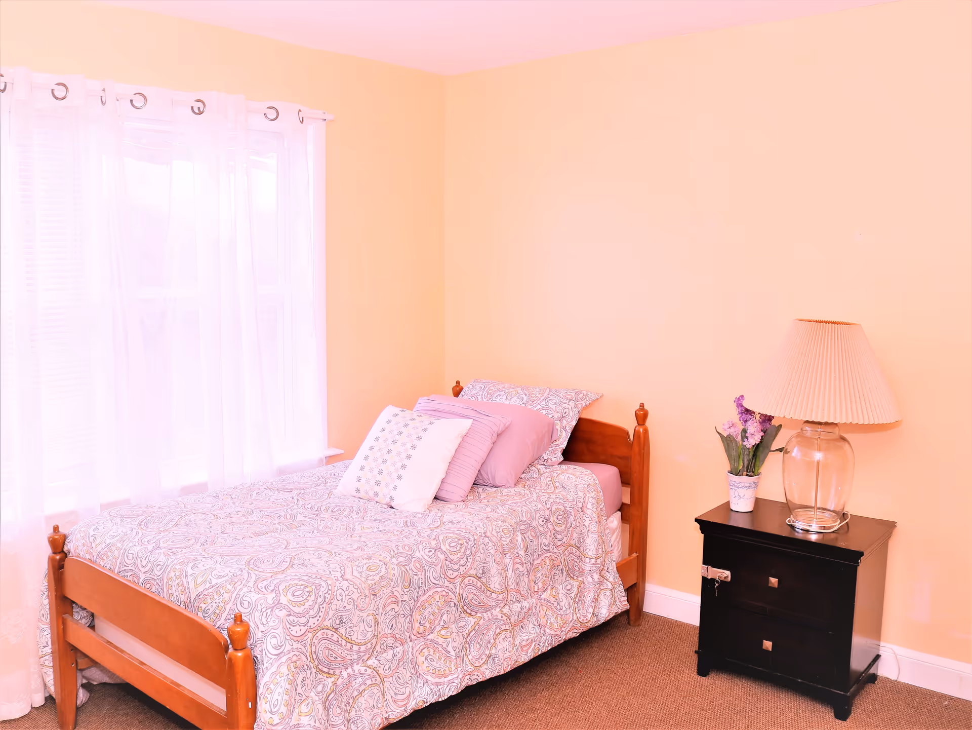 A cozy bedroom with a single wooden bed covered in a patterned bedspread and several pillows. Next to the bed is a black nightstand with two drawers, a glass lamp with a beige lampshade, and a small potted plant with purple flowers. The walls are painted light peach, and there is a window with sheer white curtains letting in natural light.