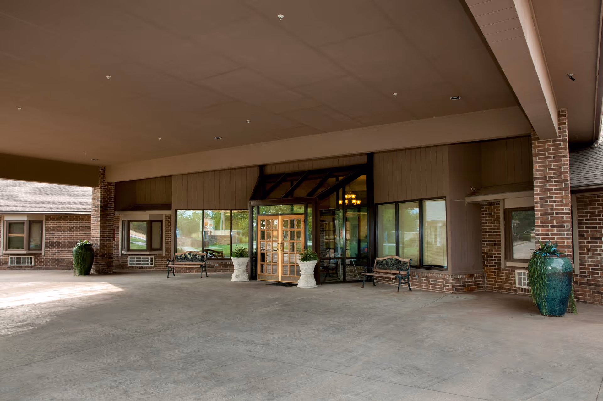 Covered entrance area of a senior living facility with a revolving glass door, two benches, large green decorative planters, and brick walls with windows.