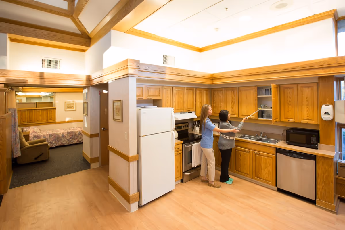 A bright kitchen area with wooden cabinets, a white refrigerator, stove, microwave, and dishwasher. Two women are standing in the kitchen; one is assisting the other in reaching into an upper cabinet using a grabber tool. In the background, there is a glimpse of a living or bedroom area with a recliner and a bed.