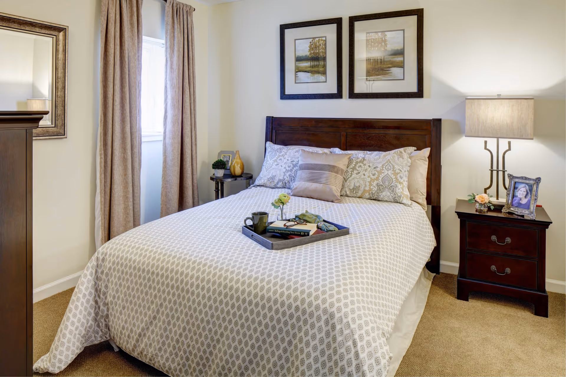 A neatly made bedroom with a wooden headboard, patterned bedding, nightstands with a lamp and framed photo, and artwork above the bed.