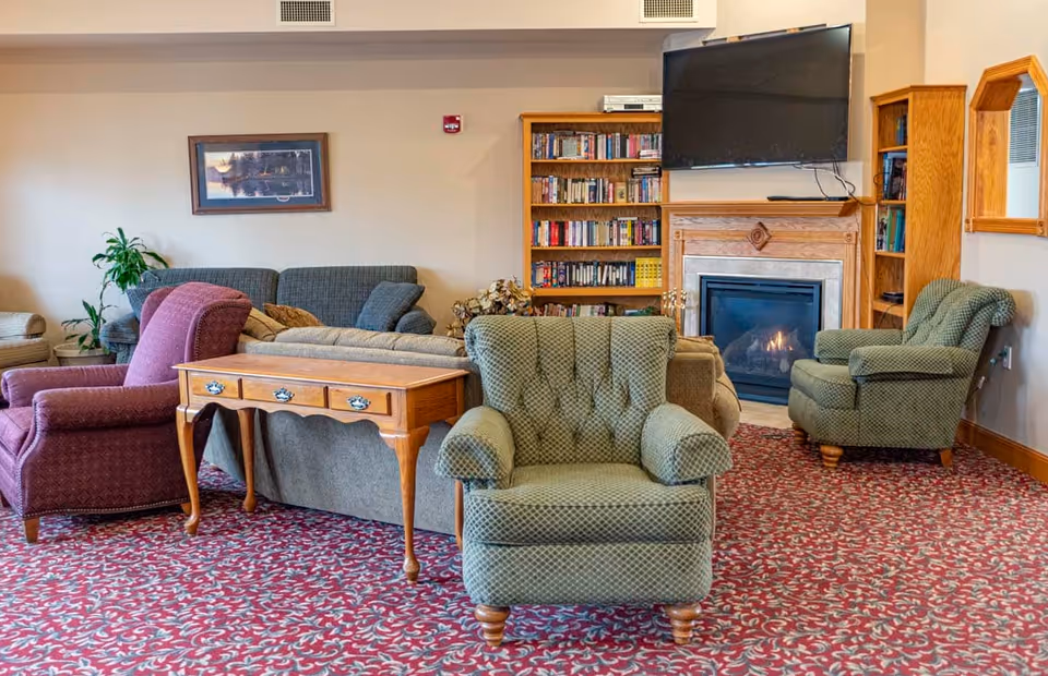 Cozy senior living common room with upholstered armchairs and sofas arranged around a fireplace, TV, and bookshelf.