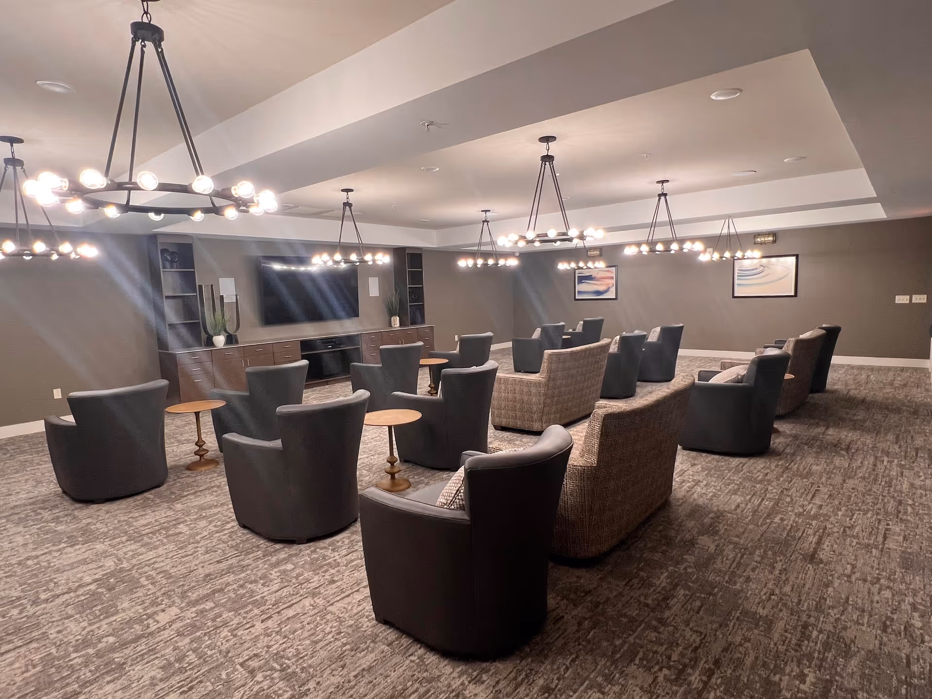 A spacious lounge area with multiple comfortable armchairs and small round wooden tables arranged in rows facing a large flat-screen TV mounted on a wall with built-in shelving and cabinets. The room has modern circular chandeliers hanging from the ceiling, gray walls, and carpeted flooring. Two framed abstract artworks are visible on the far wall.