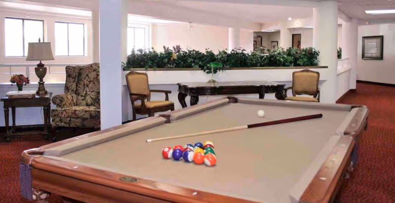 Interior view of a senior living community common area featuring a pool table with balls and cues arranged on it. The room has red carpeting, white walls, and several chairs and tables, including a floral armchair and a wooden table with a lamp and flowers. There are plants on a ledge dividing the space and framed artwork on the walls.
