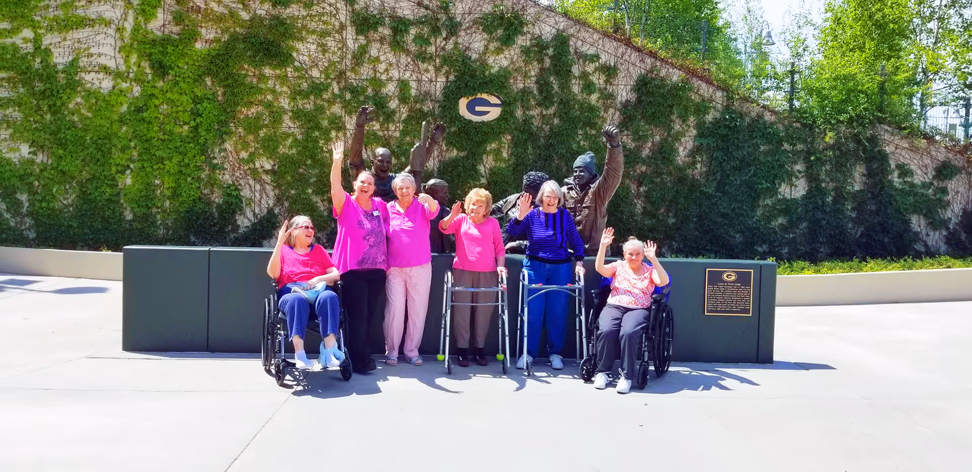 A group of elderly individuals, some in wheelchairs and others standing with walkers, posing outdoors in front of a wall covered with green ivy. They are smiling and waving, with a statue of two people raising their fists behind them. The setting is bright and sunny.