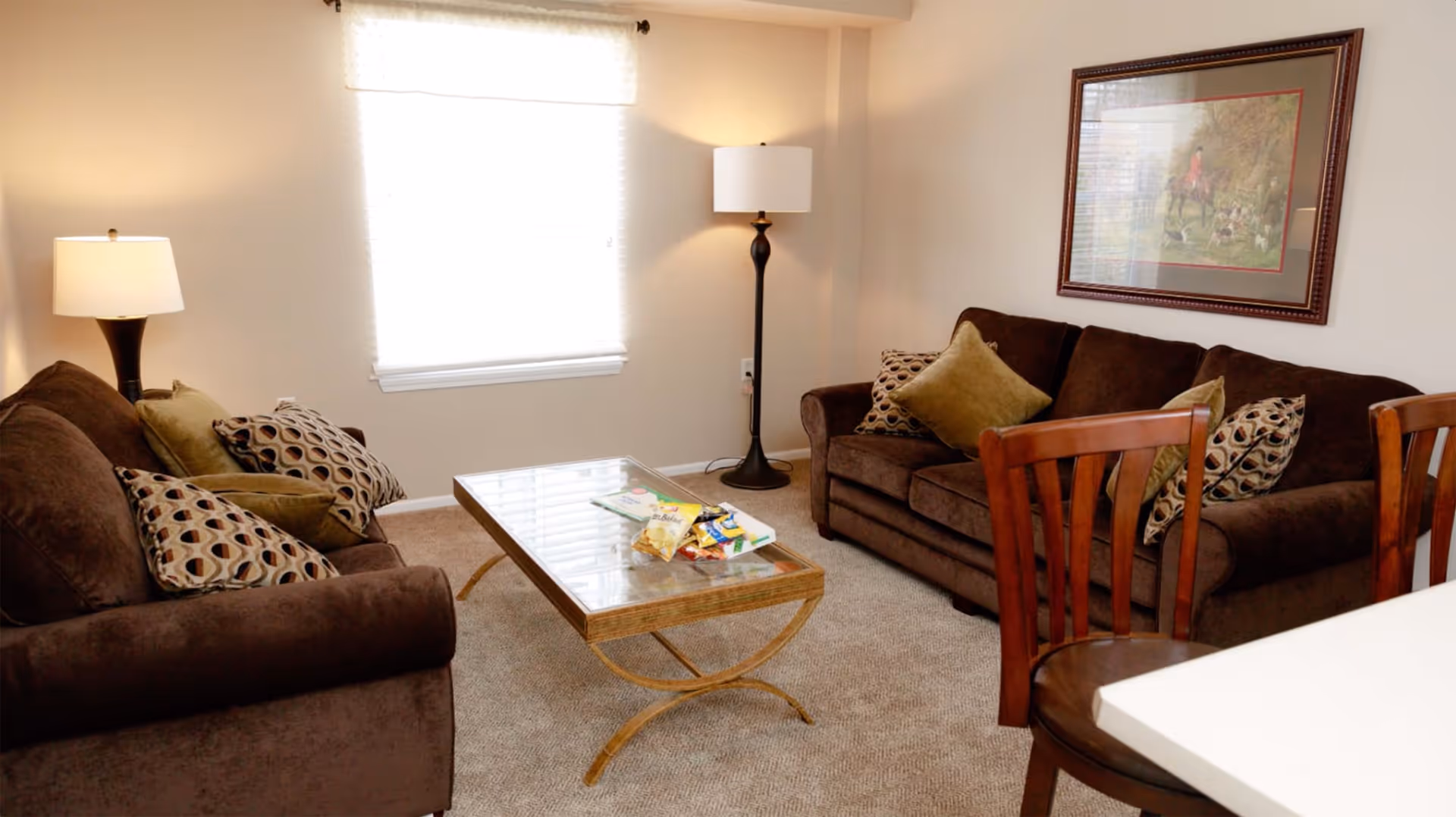 A cozy living room with two brown sofas adorned with patterned and solid pillows, a glass-top coffee table with snacks and magazines, a floor lamp in the corner, a table lamp on a side table, and a framed picture on the wall. A window with blinds allows natural light to brighten the room.