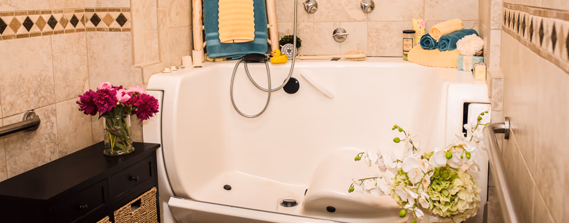 A bathroom with a white walk-in bathtub featuring built-in seats and safety handles. The walls are tiled with beige tiles and decorative diamond patterns. There are folded towels, a rubber duck, a brush, and some toiletries placed on the bathtub edges. A small black cabinet with wicker baskets and a vase of pink flowers is next to the tub, and a bouquet of white flowers is placed on the right side.