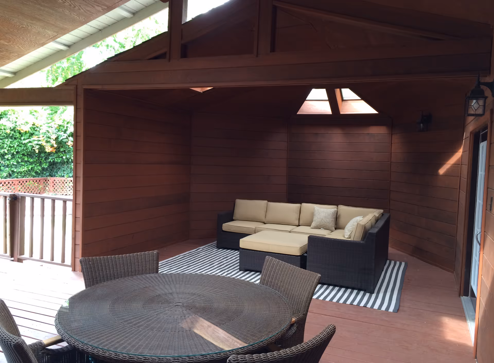 Covered outdoor patio area with wooden walls and ceiling, featuring a round wicker table with four matching chairs in the foreground and a sectional sofa with beige cushions on a striped rug in the background. Skylights in the ceiling allow natural light to enter the space.