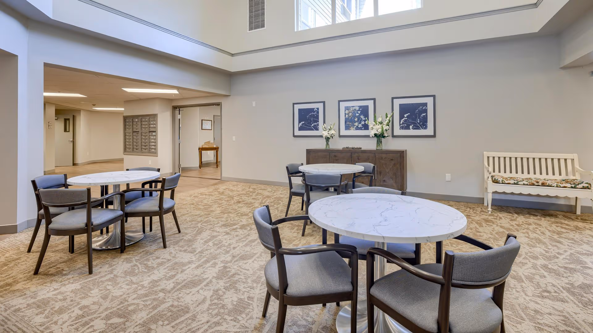 A bright, spacious interior room with round tables and chairs arranged for seating. The room has beige patterned carpet, light gray walls, and a high ceiling with large windows letting in natural light. There are three framed pictures and two vases with flowers on a wooden sideboard against the wall. A white bench with a floral cushion is placed to the right side of the room.