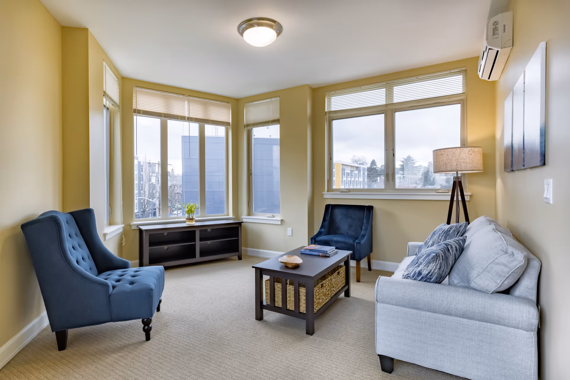A bright living room with beige walls and carpet, featuring a light gray sofa with patterned cushions, two blue upholstered armchairs, a dark wooden coffee table with woven baskets underneath, a floor lamp with a beige shade, and large windows with blinds showing an urban view outside.