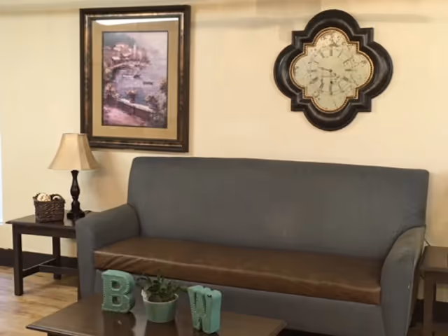 A living room area with a gray sofa that has a brown seat cushion. Above the sofa is a decorative wall clock and a framed painting of a coastal scene. On either side of the sofa are dark wooden side tables; the left table has a lamp and a basket, and the right table is empty. In front of the sofa is a small wooden coffee table with decorative letters 'B' and 'W' and a small potted plant.