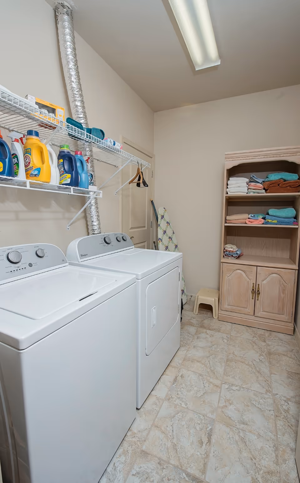 Laundry room with a washer and dryer, shelving of detergents, an ironing board, and a linen cabinet with folded towels.