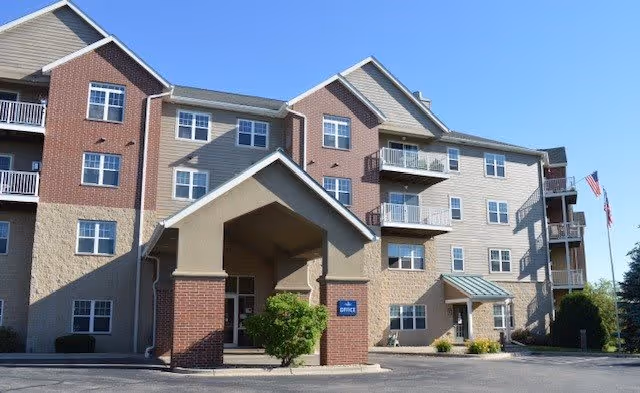 Exterior view of a multi-story senior living facility building with a covered entrance labeled 'Office'. The building has a combination of brick and beige siding, multiple windows, balconies, and an American flag on a flagpole to the right.