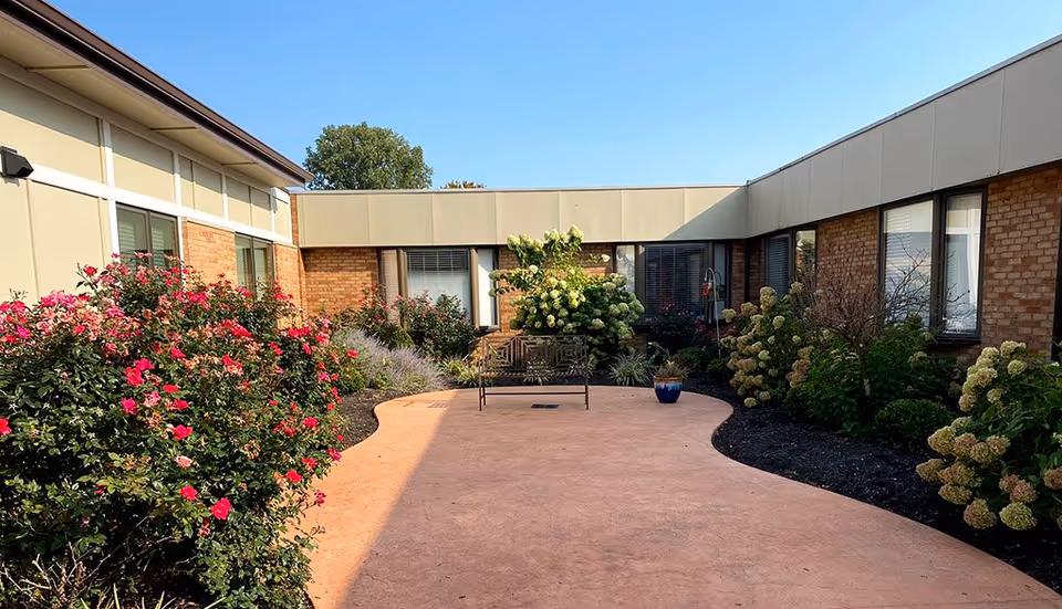 Outdoor courtyard area with a curved paved walkway surrounded by flower beds containing blooming pink and white flowers. There is a metal bench and a blue flower pot with plants in the center of the courtyard. The courtyard is enclosed by a single-story building with windows and brick walls under a clear blue sky.