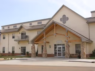 Exterior view of a senior living facility building with a covered entrance supported by stone pillars, beige siding, and multiple windows including a small balcony.
