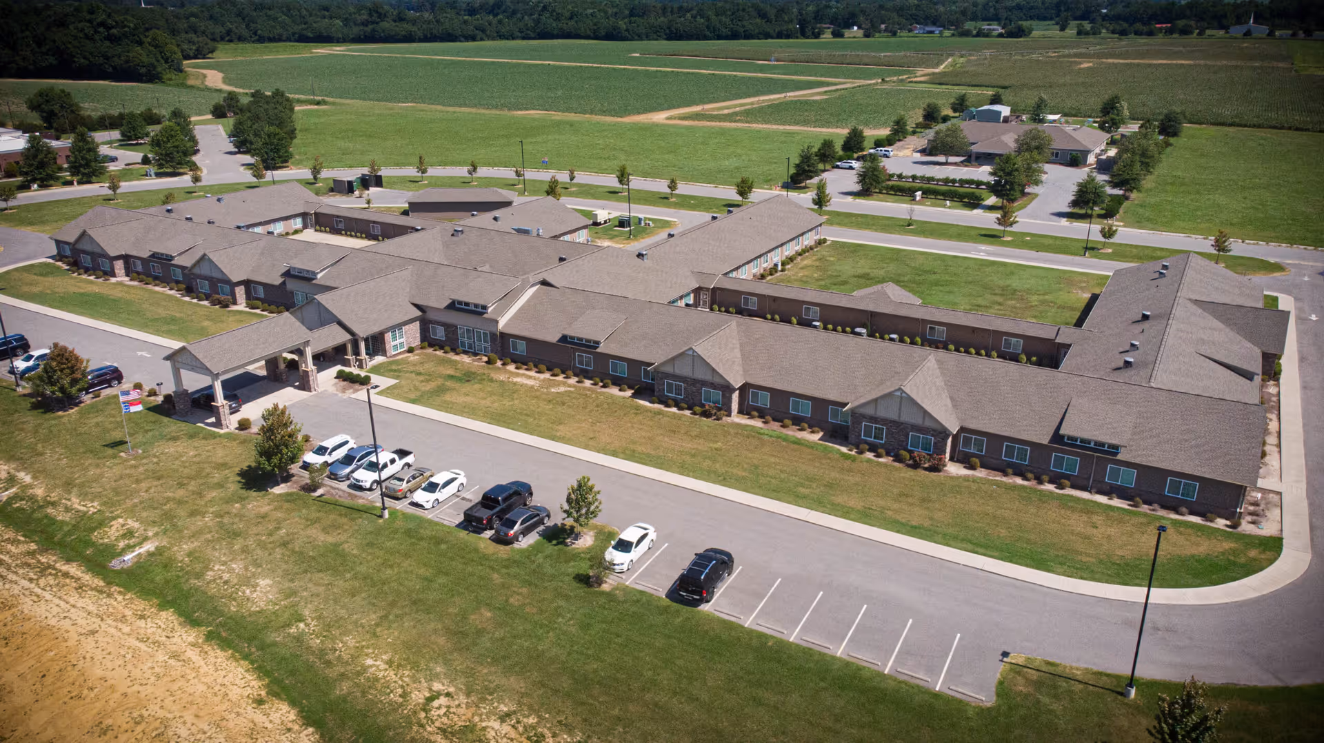 Aerial view of a single-story senior living complex with a covered entrance, parking lot, lawns, and surrounding farmland.