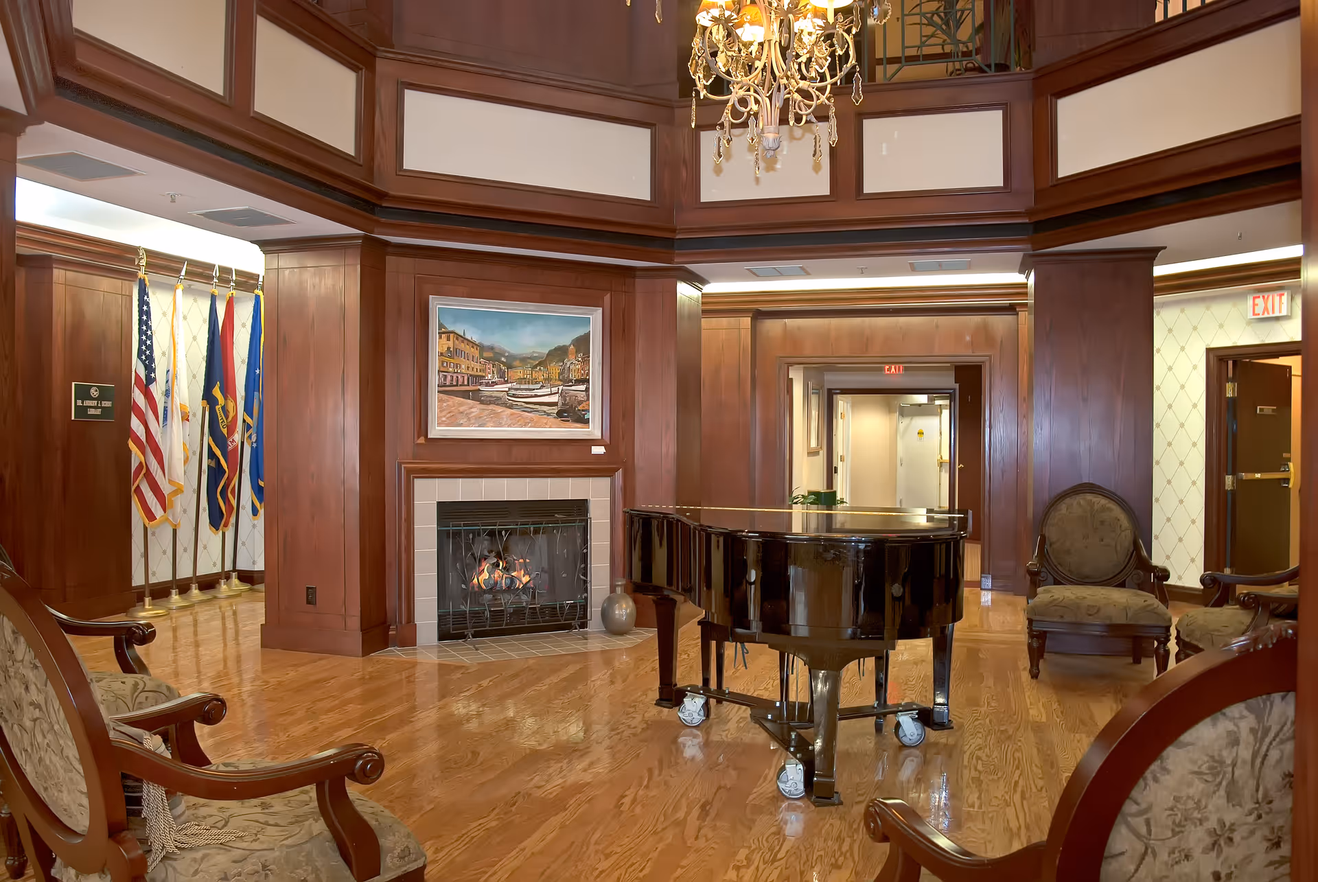 A spacious, elegant living room area with polished wooden floors and wood-paneled walls. The room features a black grand piano on wheels in the center, a lit fireplace with a painting above it, several upholstered chairs with wooden frames, and a chandelier hanging from the ceiling. Flags are displayed in the corner near a plaque, and there are exit signs visible above doorways.