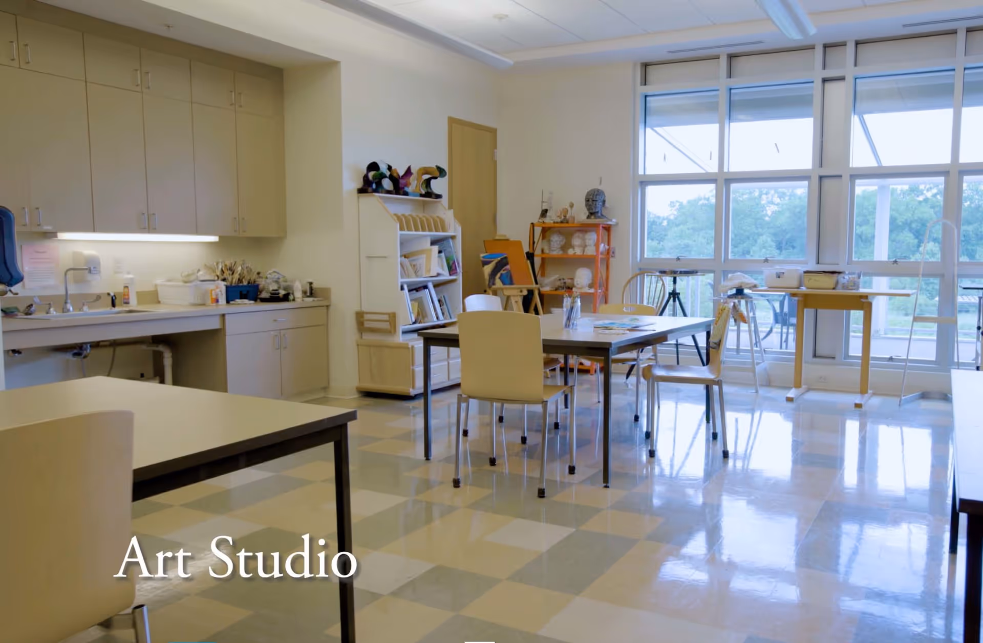 A bright art studio room with tables and chairs arranged for creative activities. There are shelves with art supplies, a sink with cabinets, and large windows letting in natural light with a view of greenery outside.
