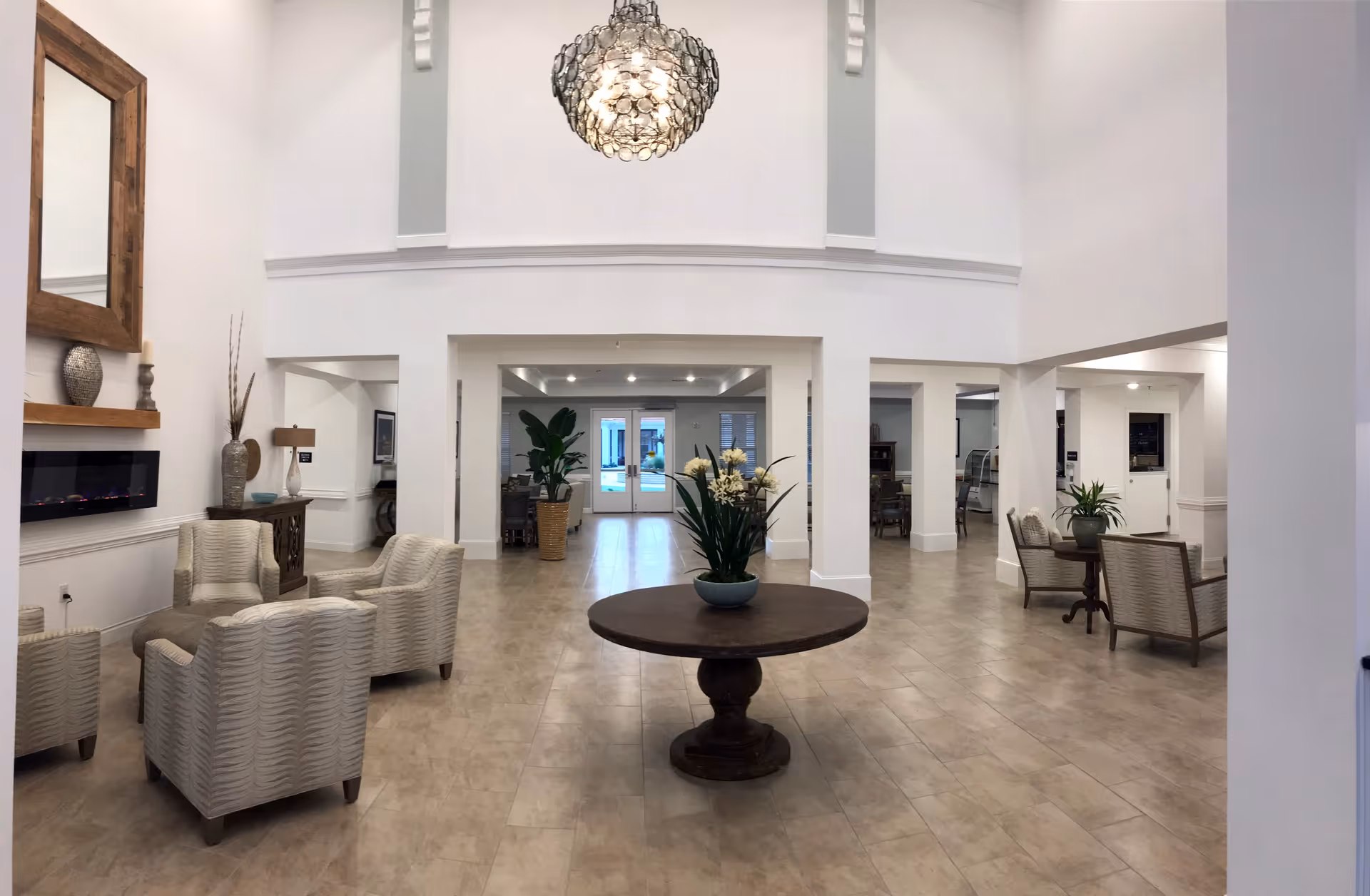 Spacious and well-lit senior living facility common area with beige tiled flooring, white walls, and a high ceiling. The room features a round wooden table with a flower arrangement in the center, several beige upholstered armchairs arranged in small seating groups, a large decorative mirror above a modern electric fireplace, and potted plants. A large chandelier hangs from the ceiling, and double glass doors are visible at the far end of the room.