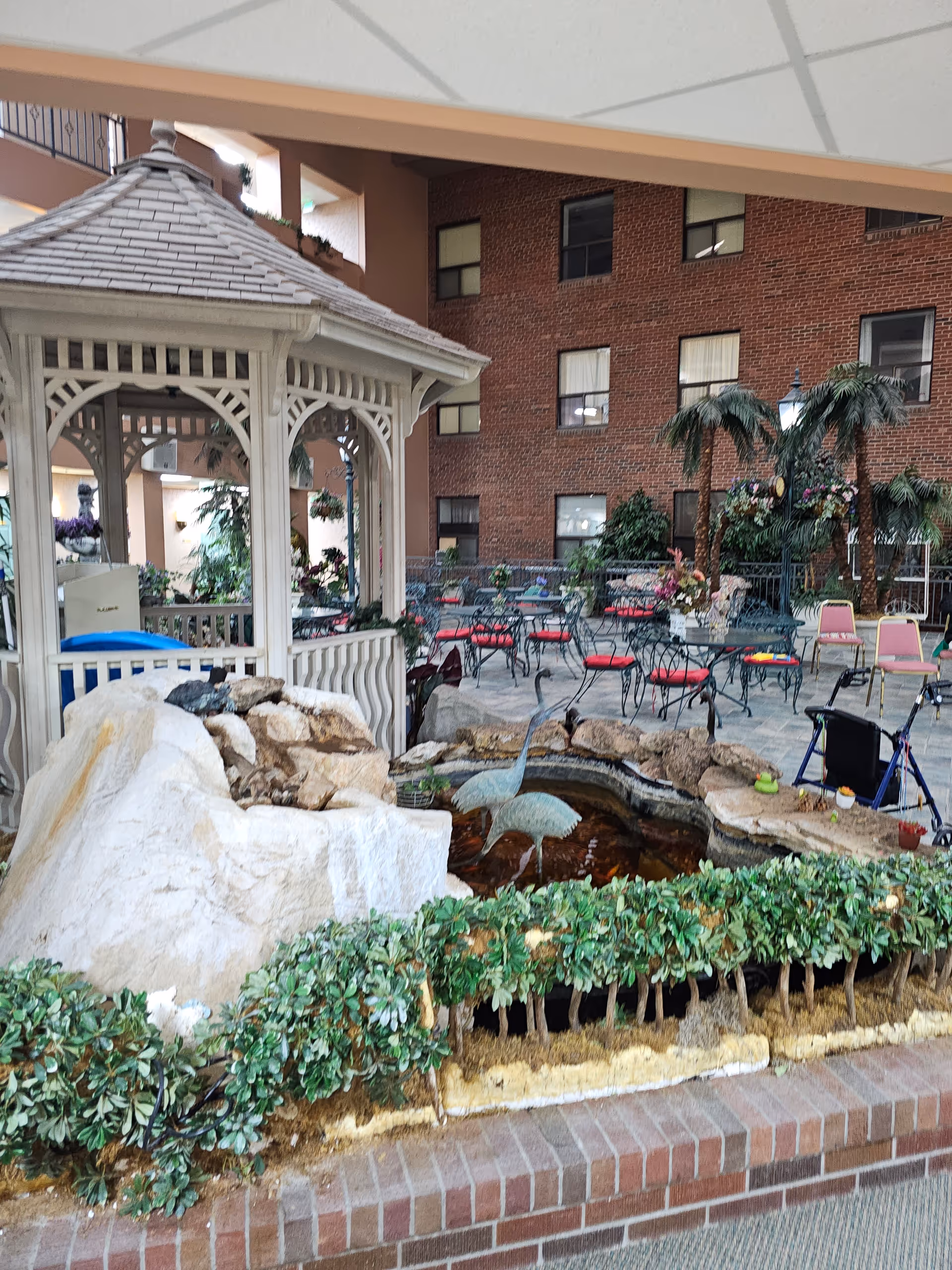 Indoor courtyard area with a white gazebo, a small pond featuring two metal bird sculptures, surrounded by rocks and greenery. In the background, there are several tables and chairs with red cushions, potted plants, and a brick wall with windows. A walker is also visible near the seating area.