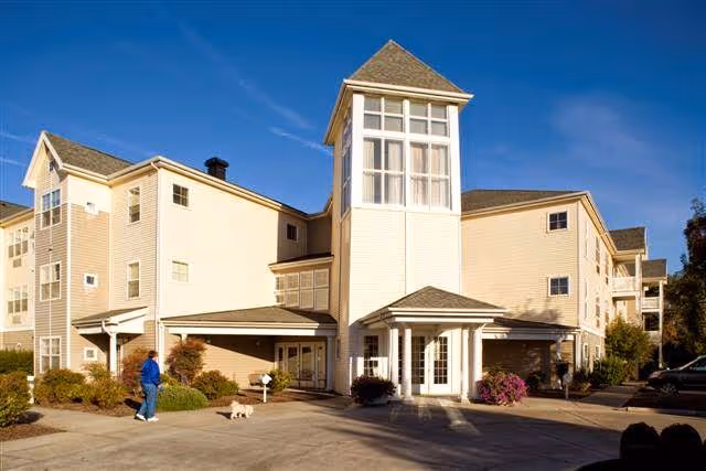 Exterior view of a multi-story senior living facility building with beige siding and a prominent tower-like entrance. A person in a blue jacket is walking a small dog on the driveway in front of the building. The sky is clear and blue.