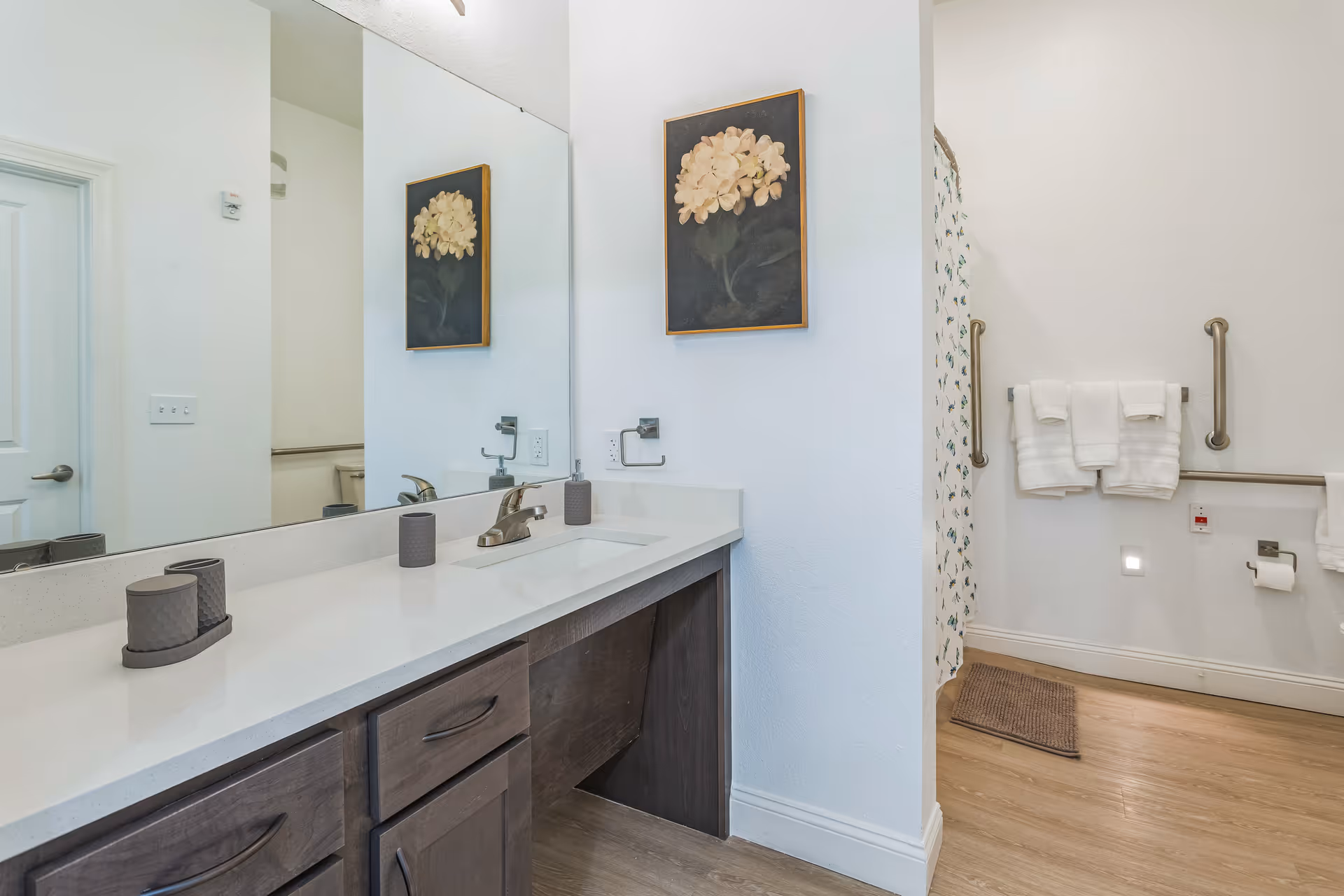 A clean and modern bathroom featuring a long white countertop with a built-in sink, dark wood cabinets underneath, a large mirror above the sink, and a wall-mounted floral painting. The bathroom has a shower area with a floral shower curtain, grab bars, and neatly folded white towels hanging on the wall. The floor is light wood, and the walls are painted white.