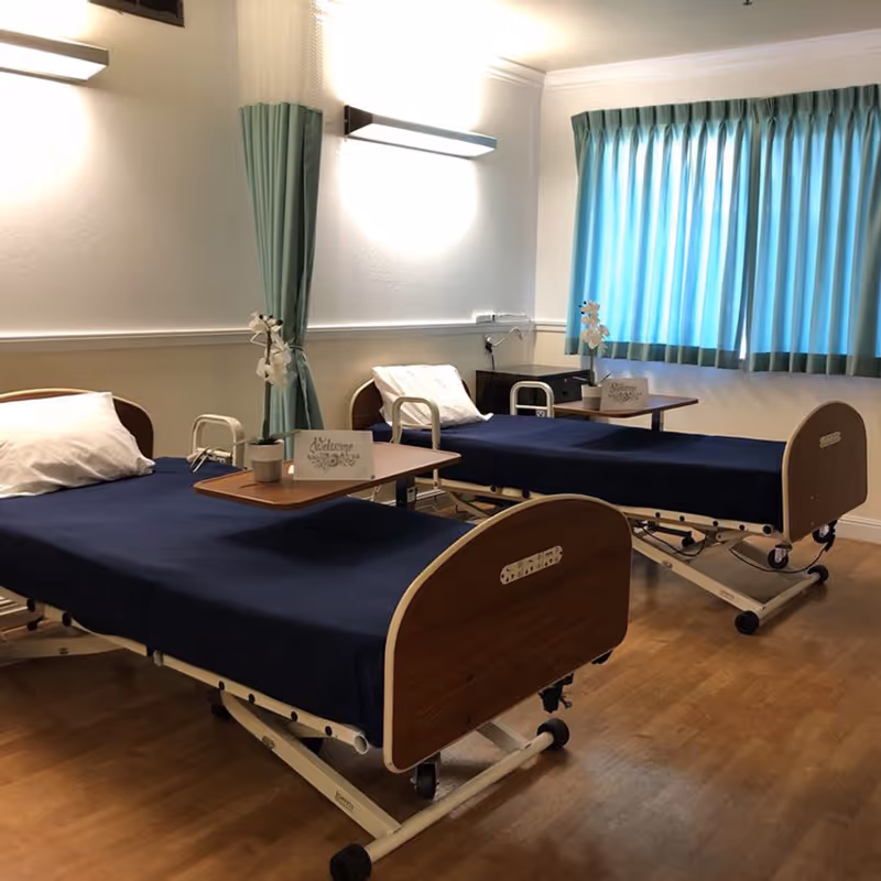 A room with two hospital-style beds with navy blue bedding and white pillows. Each bed has a wooden overbed table with a small potted plant and a welcome card. The room has light-colored walls, wooden flooring, a window with blue curtains, and wall-mounted lights above each bed.