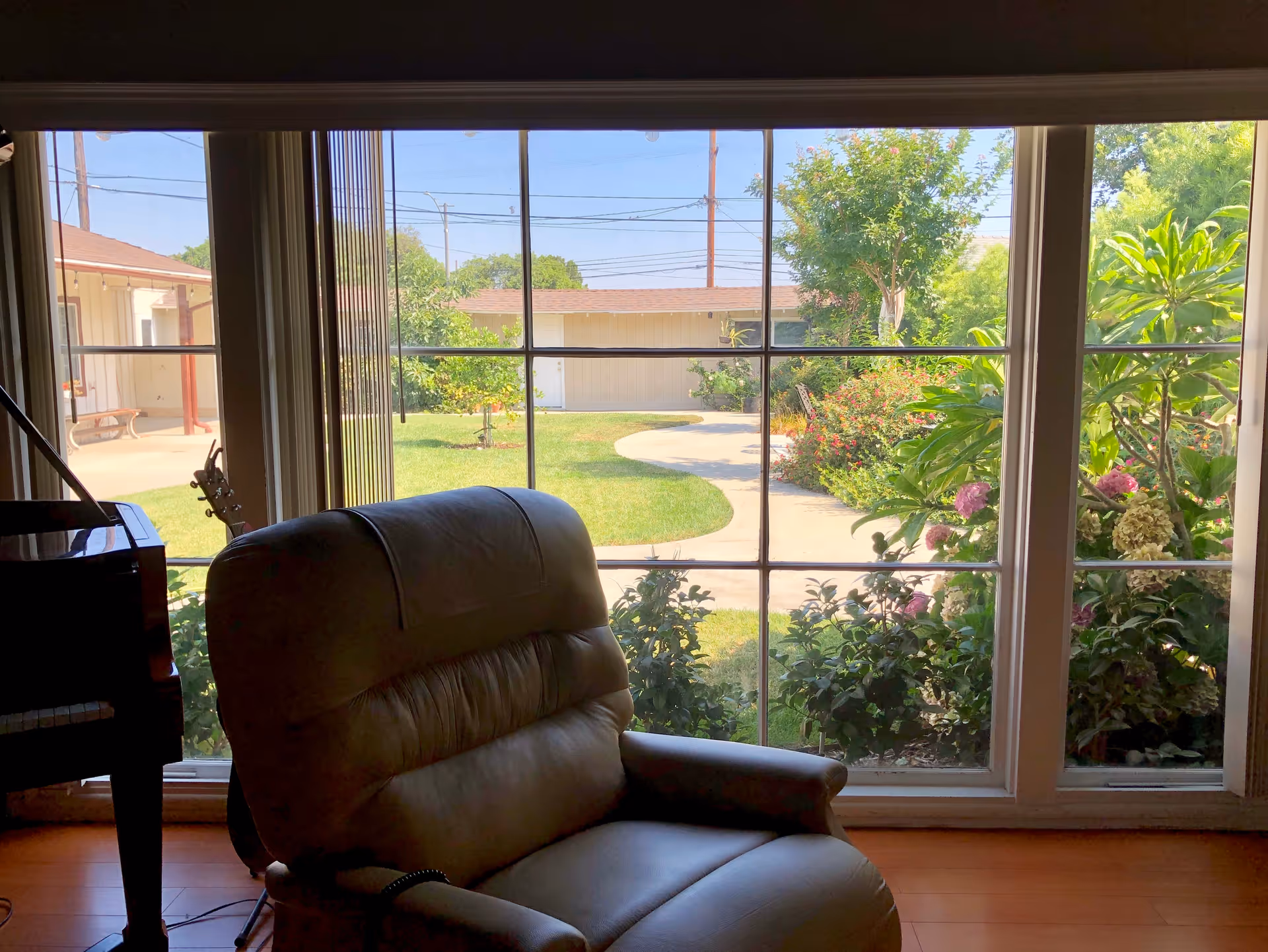 A cozy leather armchair positioned near a large window with a view of a garden path, green bushes, and trees outside. Part of a piano and a guitar are visible on the left side inside the room.