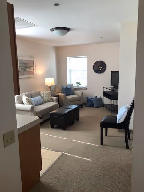 A cozy living room with a beige sofa and armchair, both adorned with decorative pillows. There are two black ottomans in front of the sofa, a black chair with a pillow on the right, and a small TV on a black stand against the wall. A round wall clock hangs above the TV, and a window with a small plant on the sill lets in natural light. A floor lamp and framed artwork are visible on the left wall.