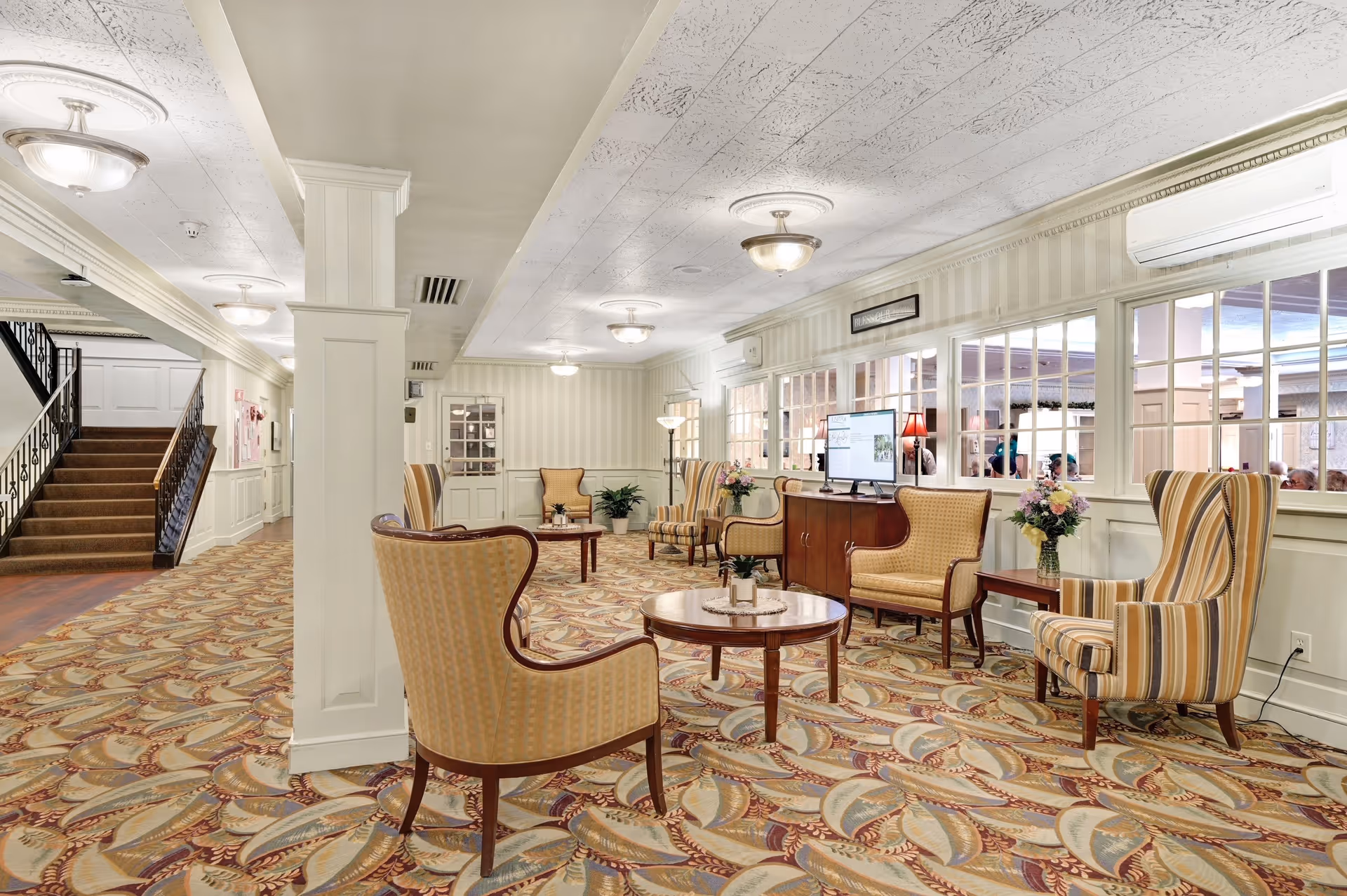 A spacious, well-lit common area in a senior living facility featuring patterned carpet, several upholstered armchairs and small wooden tables arranged for conversation, a wooden cabinet with a computer monitor on top, and large windows looking into another room. The area has cream-colored walls with vertical stripes, ceiling lights, and a staircase leading to an upper floor.